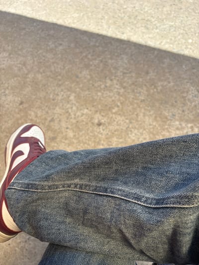The image shows a close-up of a person's legs, wearing blue denim jeans and a maroon and white sneaker, resting on a concrete surface. The concrete appears weathered and has a shadow cast across it. The background is out of focus. The scene is not identifiable as being in Nukus, Uzbekistan or any specific city.