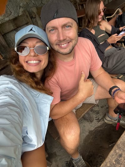 A group of people are resting in an outdoor setting. A man wears a gray cap and a pink shirt, while a woman sports a light blue cap and circular sunglasses. The woman wears a light blue shirt and is smiling. Another person is seen partially in the background. The setting appears to be a rest stop along stone walkways. Location: possibly in or around Yên Minh Commune, Vietnam.