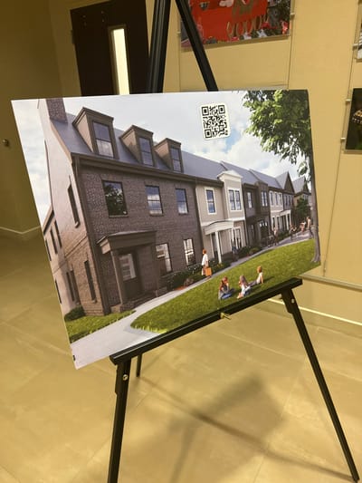 A printed image of multi-story townhouses is on an easel. The buildings have a brick exterior with numerous windows and dormers. A strip of green grass and a paved sidewalk appear in front of the townhouses. Figures of people are digitally added to the grass, suggesting a residential community. A QR code is positioned in the top left corner of the image, likely linking to additional information. The picture is angled and slightly out of focus, with the easel and surrounding background visible.