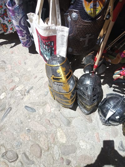 The image shows a street-level view of items for sale, likely in a market or shop, on a cobblestone surface in Besalú, Spain. In the foreground, there are three decorative knight's helmets, two standing upright and one on its side. The helmet on the left is dark gray with gold trim and has a segmented visor. The helmet in the middle is also dark gray with gold accents on the faceplate, featuring intricate detailing. The helmet on the right is silver and partially visible, appearing to be in a similar style. Behind the helmets, there are several tote bags. One white canvas bag with red trim and an illustration of a building and plants is prominent. To its left, another tote bag with a patterned design of purple flowers and a lizard is visible. Further back, there is what appears to be a child's costume armor, made of a mesh-like material with metallic accents, and a collection of wooden toy weapons, including staffs and possibly a toy axe. The ground is made of irregular cobblestones of various colors, including gray, pinkish-brown, and off-white, with visible gaps filled with sand or dirt. The lighting suggests it is daytime, with strong shadows cast by the objects and an unseen person.