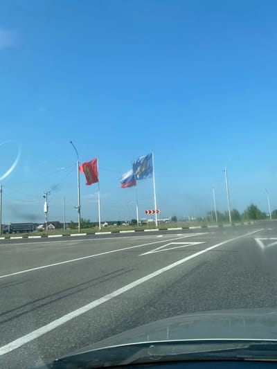 The image shows a road in Селиваниха, Russia, on a clear day. The sky is blue with a few wispy clouds. On the left side of the road, there are two flags flying on tall poles. The flag on the left is red with a gold emblem. The flag on the right is blue and white with a gold eagle emblem. The Russian flag is also visible to the right of the red flag. In the background, there are some houses and trees. The road has white lines marking the lanes and arrows indicating traffic direction. A truck is visible on the left side of the road. The image appears to be taken from a vehicle.
