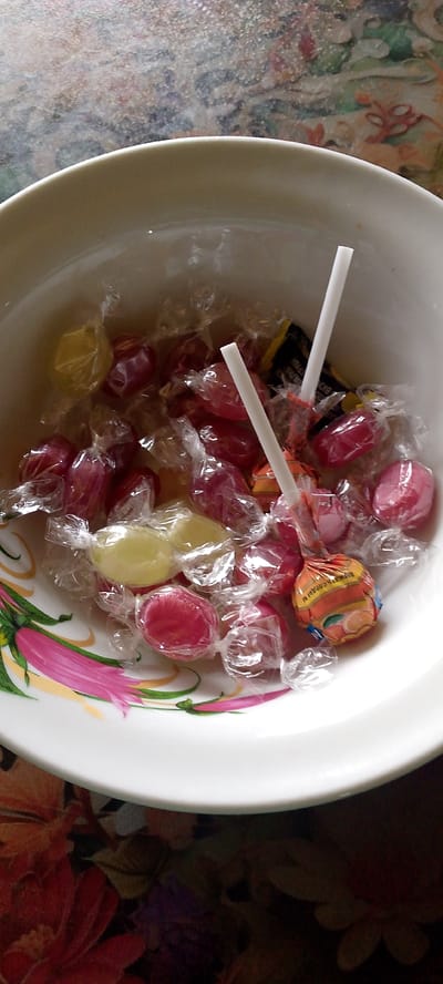 A white ceramic bowl filled with individually wrapped hard candies is placed on a surface with a floral pattern. Two white lollipop sticks emerge from the candies. The bowl has a decorative rim with pink and green floral designs. The candies appear to be various colors, including red, yellow, and orange, and are wrapped in clear cellophane. One candy wrapper, orange with yellow and red stripes, is partially visible, showing Cyrillic text. The background surface has a busy, colorful floral print. The overall scene suggests a display of sweets. The city of Shkurinskaya, Russia, is not depicted in this image.