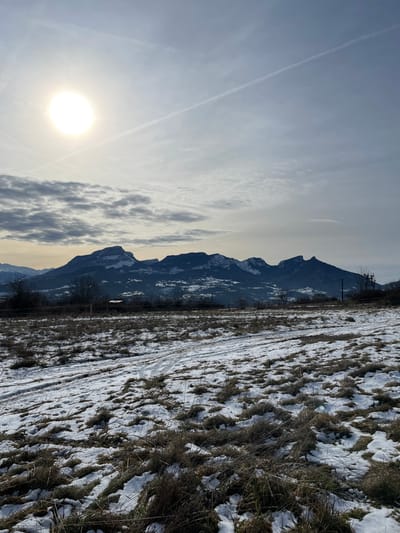 The image depicts a winter landscape in or near Chambéry, France. The foreground consists of a field with sparse, dry brown and green vegetation interspersed with patches of white snow, showing visible tire tracks or paths in the snow and exposed earth. In the middle ground, a horizontal fence or barrier stretches across the scene, backed by a line of leafless trees. To the far right, a utility pole with attached electrical apparatuses is visible. The background features a prominent range of dark, jagged mountains, several peaks of which are capped with snow. The sky above is partially cloudy, predominantly light grey, with a bright, circular sun positioned in the upper left quadrant. A distinct white airplane contrail extends diagonally from the upper left to the upper right across the sky. No people or animals are present.
