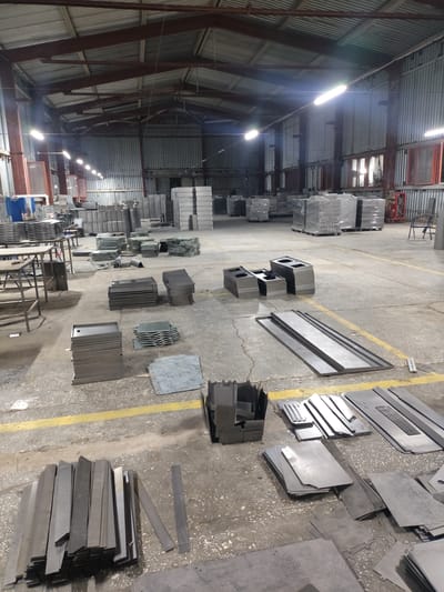 The image depicts the interior of a large industrial warehouse, likely a metal fabrication facility, located in Dangir, Uzbekistan. The space is cavernous with a high, corrugated metal roof supported by exposed metal beams and columns. Fluorescent lights are suspended from the ceiling, illuminating the concrete floor. The floor is largely empty but is covered with numerous stacks of metal components, sheets, and fabricated parts.
In the foreground, a large pile of long, flat metal strips is arranged horizontally. To the left and slightly behind this, a stack of rectangular metal pieces with holes and cutouts is visible. Further back, various other stacks of metal parts are scattered across the floor, some neatly arranged in piles and others more haphazardly. There are also several large, flat metal sheets laid out.
In the middle ground, there are tables with metal structures, possibly workbenches or assembly stations. Some of these tables have metal components on them. In the background, more stacks of metal are visible, including some large, wrapped pallets. The walls of the warehouse appear to be constructed from corrugated metal siding, with windows visible in some sections. The overall impression is one of a busy manufacturing environment with a large volume of metal stock and fabricated items. No people are clearly visible in the image, suggesting the space may be photographed during a break or at the end of a workday.