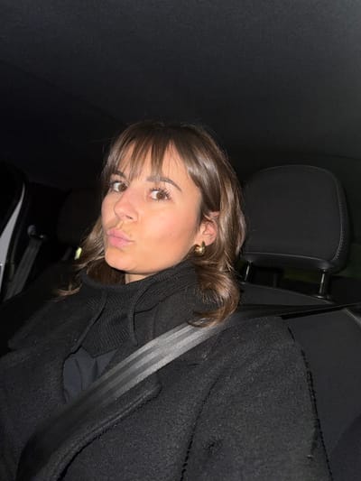 A young woman with brown hair and bangs poses in the passenger seat of a car. She has brown eyes, fair skin, and is wearing gold hoop earrings. A black jacket and a seatbelt obscure part of her body. The car interior is dark, with black seats and a headrest visible. Her lips are pursed in a slight pout. The image is rotated, giving a tilted perspective.