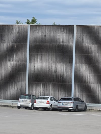 The image shows a row of parked cars against a wooden slatted wall. The cars are a silver Volkswagen Passat, a white hatchback, and a silver small hatchback. The Passat has the license plate "WI-MH 56". The white hatchback has the license plate "JB-5796". The silver small hatchback has the license plate "SG-BN 209". The wooden wall appears to be a sound barrier, common in areas near roads or industrial zones. The sky is visible on the left side of the image, reflecting in a metallic surface which may be part of the wall or a separate structure. Green foliage from trees is also visible, some of which appears to be reflected. The scene suggests an outdoor setting, likely a parking lot or a roadside. The lighting indicates it is daytime, with clear skies. The environment appears to be urban or suburban, given the presence of cars and the sound barrier. There are no people visible in the image.
