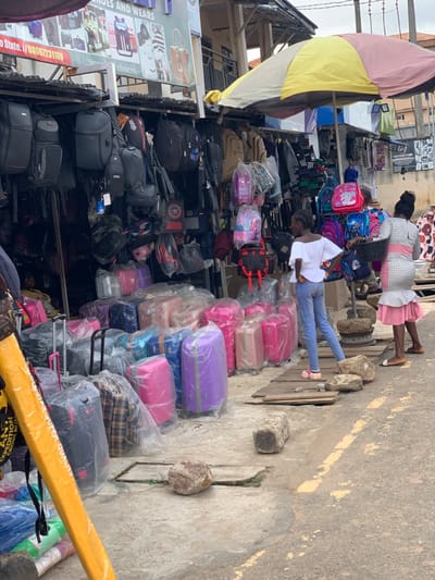 The scene is a street market in Ondo, Nigeria, located at 7.252 latitude, 5.199 longitude. There are many colorful suitcases on display, some wrapped in plastic, as well as backpacks and other luggage. Three people are present: a woman in a pink dress, a woman in a white shirt and blue jeans, and a woman in a white shirt and black pants. The woman in the pink dress is standing near the suitcases, looking at the camera. The woman in the white shirt and blue jeans is standing near the backpacks, looking to the left. The woman in the white shirt and black pants is standing behind the woman in the pink dress, looking to the right. The market is set up in front of a building with a sign that reads “SHOES AND WEARS.” There is a yellow umbrella above the market, and the ground is concrete.