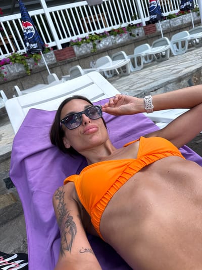 In the city of Grivitsa, Bulgaria, a woman is lying on a lounge chair covered by a purple towel. She is wearing an orange bikini and black sunglasses. Her left arm is resting on the back of the chair, and she is holding her sunglasses with her right hand. The background shows a row of white lounge chairs and umbrellas with the Pepsi logo. The scene appears to be outdoors, possibly at a resort or a pool area, with lush greenery and stone landscaping.