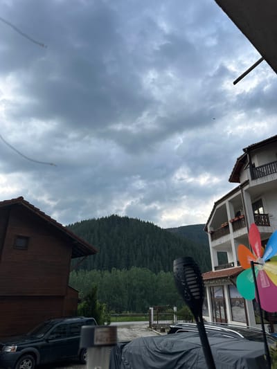 The image captures a rural landscape in Madzhare, Bulgaria, under an overcast sky. The scene is dominated by natural elements in the background and architectural structures with vehicles in the foreground.

The sky is filled with dense, uniform gray clouds, suggesting a cloudy day, with some lighter patches visible. Two faint, curved streaks, possibly contrails, are present in the upper left quadrant of the sky. A small, dark, winged object is visible flying in the upper right sky.

In the background, a large, densely forested mountain range rises prominently, covered by coniferous trees. Below the main forest, a band of lighter green deciduous trees is visible, with hints of a road or path partially obscured by vegetation to the far right.

On the left side of the foreground, a two-story wooden house with a reddish-brown tiled roof is positioned. It features horizontal wooden siding and a small, square window on its upper floor. A dark green first-generation Land Rover Freelander SUV is parked in front of this wooden house, facing left. To the right of the Land Rover, partially obscured by an object in the immediate foreground, another vehicle is visible, covered with a dark gray tarp. A small, conical evergreen tree stands between the wooden house and the paved area.

On the right side of the foreground, a multi-story white building with a reddish-brown tiled roof is present. This building features multiple balconies with dark wooden railings and visible flower boxes. The ground floor of this building includes large glass windows or doors that show reflections. A colorful pinwheel with red, orange, green, blue, and pink blades is prominently positioned in the lower right corner of the frame, partially in front of the white building. A black, torch-shaped outdoor light fixture, designed to mimic a flame, rises from the bottom center of the image, partially obscuring the tarp-covered vehicle and the pinwheel. The ground in the immediate foreground is a paved surface, likely a driveway or parking area. No people are visible in the image.