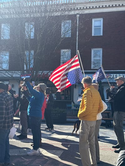 A small group of people stand on a street, several holding signs. An American flag waves prominently. A brick building is visible behind the crowd with several windows. Some people wear casual clothing, including a yellow jacket and a blue jacket. A dog is also present in the scene. Sunlight casts shadows across the street.