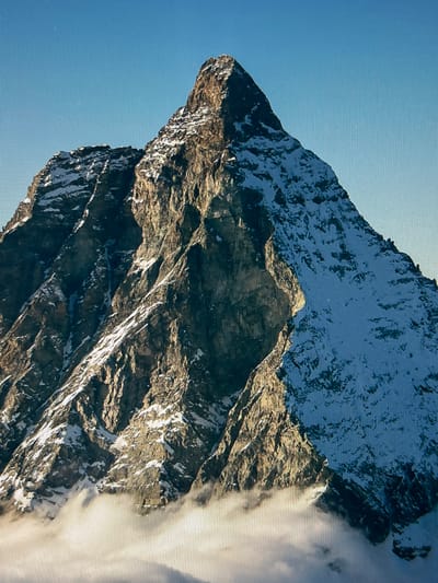 This is a media file depicting a dramatic mountain landscape under a clear blue sky. The primary subjects are the rugged, rocky slopes of a mountain, partially covered in snow. The mountain is characterized by sharp angles and steep inclines, suggesting a formidable and imposing presence. The texture of the rock is rough and appears weathered, with areas of light and shadow emphasizing its topographical features. Patches of snow cling to the higher elevations and crevices, contrasting with the darker rock.

In the foreground and to the right of the frame, a bank of soft, white clouds partially obscures the mountain, creating a sense of depth and atmosphere. The clouds appear to be swirling around the peaks, hinting at the dynamic weather conditions at high altitudes. The overall scene conveys a sense of grandeur and the raw power of nature.

The lighting suggests that it is daytime, likely late morning or early afternoon, given the strong sunlight illuminating the mountain slopes and casting deep shadows. The clear blue sky indicates fair weather, though the presence of clouds at the mountain's elevation suggests a cooler climate. There are no people or man-made objects visible in the image, emphasizing the wild and untamed nature of the setting. The image has a slightly grainy texture, possibly due to the camera or display technology. There is no visible text in the media file.