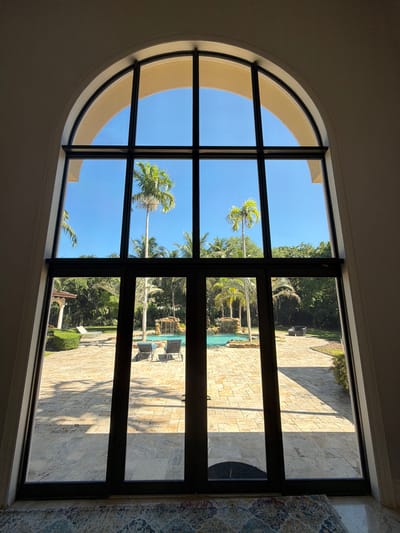 The image shows a large arched window with black framing, looking out onto a backyard oasis. The backyard features a swimming pool with a waterfall, surrounded by lush tropical foliage and palm trees. There are lounge chairs by the pool and a paved patio area. The sky is a clear blue, suggesting a sunny day. The window itself is large and imposing, with multiple panes of glass that divide the view of the outdoors. The interior of the room where the photo is taken is dimly lit, with a patterned rug visible on the floor in the foreground. This scene is consistent with a luxurious residential property in Pinecrest, Florida, a city known for its sprawling estates and tropical landscaping.