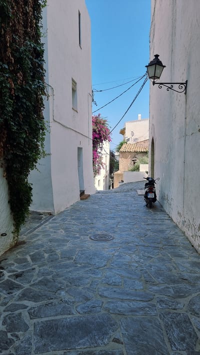 This image captures a narrow, cobblestone alleyway in Cadaqués, Spain, on a clear day. The perspective is low and tilted, looking up the lane. On the left, a whitewashed building with a dark green, leafy vine climbing its lower section dominates the foreground. Above this, a vintage black lamppost with ornate scrollwork is affixed to the wall. Further up the alley, a bright pink bougainvillea bush adds a splash of vibrant color against the white buildings and the clear blue sky.
On the right side of the alley, the stone pavement is laid with irregular grey flagstones. Partway up the lane, a black scooter is parked facing away from the camera. Its license plate is visible, with text that appears to be "BCE 3804". The scooter is the only sign of modern activity in an otherwise historic-feeling scene.
The architecture suggests a Mediterranean setting, characteristic of coastal towns like Cadaqués. The bright sunlight indicates it is daytime, likely late morning or afternoon given the shadows. The overall mood is tranquil and picturesque, evoking a sense of timeless charm. The scene is devoid of people, emphasizing the quiet solitude of the alley.