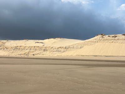 The image depicts a coastal landscape with large sand dunes under a dramatic sky. In the foreground, a sandy beach stretches out, marked with faint footprints and some scattered debris. A shallow, wet sand area runs horizontally across the mid-ground, separating the beach from the base of the dunes. The dunes themselves are prominent, characterized by layered striations and a weathered, textured appearance. They rise to a crest that forms the horizon line. Above the dunes, a dark, ominous cloud bank dominates the upper portion of the sky, contrasting with a lighter, partly cloudy sky to the right. Several small figures are visible at the base of the dunes, appearing to be walking or interacting. Based on the context provided, this scene is located in the city of Lège-Cap-Ferret, France, likely near the Dune du Pilat, known for its vast sand dunes and coastal environment.