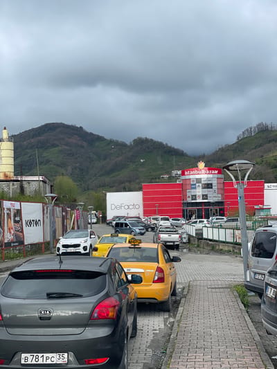 The image shows a parking area in Kemalpaşa, Turkey. There is a Kia vehicle in the foreground, with the license plate B781PC 35 RUS. Various other cars, including yellow taxis, are lined up. In the background, a large building is branded with "istanbulbazaar" and other brand names, including LC Waikiki and DeFacto. The surrounding environment features a hillside and overcast sky. There are various commercial banners for Koton and Flo, along with other objects like light posts and fences.