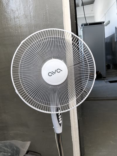 An indoor shot shows a white pedestal fan with the brand name "ava" visible on its central hub. The fan is positioned in front of a grey, textured wall. To the right of the fan, a doorway leads to an area with industrial-looking equipment, including a grey metal cabinet and some electrical wiring. The floor in the background appears to be concrete. In the lower left corner, a clear plastic bag containing dark items is visible. The scene suggests an interior space, possibly a workshop or storage area.