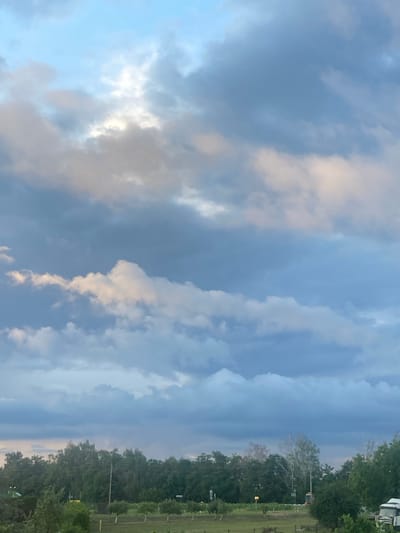 The image captures a view of the sky over a rural landscape, likely in Матыкальскі сельскі Савет, Belarus. The sky is filled with a mix of clouds, ranging from light, wispy formations to heavier, darker masses, indicating potentially dynamic weather conditions. The upper portion of the sky is a soft blue, transitioning to grayer tones as clouds dominate. Sunlight is breaking through some of the clouds, creating brighter patches. Below the sky, a dense line of green trees forms the horizon, with various types of foliage visible. In the foreground, there are hints of agricultural fields or parkland with manicured areas and some scattered trees. The overall impression is of a serene, natural environment during a period of changing weather.