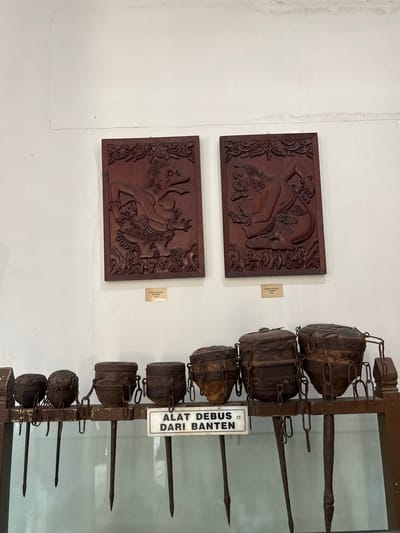 The image depicts a display within a museum, likely the Museum Pusaka Keraton Kasepuhan in Cirebon, Indonesia, given the presence of Wayang reliefs and traditional tools. Two wooden reliefs of Wayang figures, possibly characters from Javanese puppetry, are mounted on a white wall. Below these reliefs, a collection of what appear to be historical ritualistic or ceremonial tools is displayed on a wooden shelf behind glass. These tools consist of several cylindrical, metal containers with chain attachments, each suspended by a long, pointed metal stake. A white sign with black text in Indonesian, "ALAT DEBUS DARI BANTEN," is placed in front of the tools, identifying them as "Debus tools from Banten." The overall setting suggests an indoor exhibition space with neutral-colored walls and a display cabinet.