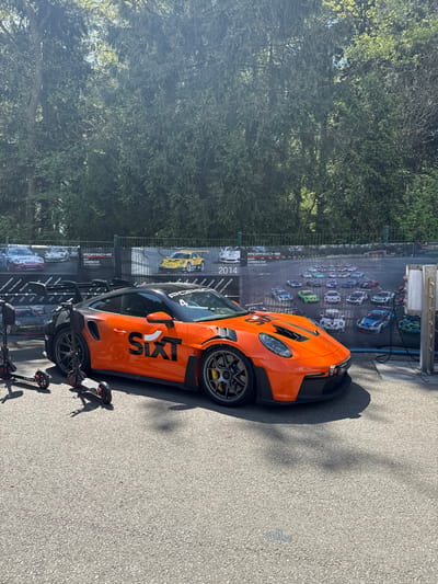 An orange Porsche 911 GT3 Cup car with the number 4 on its windshield and "SIXT" branding on its side is parked on a gray asphalt surface in front of a large white and green billboard. The billboard features photographs of multiple Porsche race cars and the words "PORSCHE" and "SIKT" in black. The car is facing the camera and its front end is slightly angled to the left. There are two black electric scooters parked to the left of the car. The scene is set against a backdrop of a dense, green, leafy forest. The location is in the town of Stavelot, Belgium, at the coordinates 50.441 latitude and 5.969 longitude.