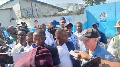 The scene depicts a dense gathering of approximately 15-20 male individuals outdoors in the city of Kabinzi, Rwanda, under bright daylight. The majority of the individuals are dark-skinned, with one light-skinned male visible in the foreground. This light-skinned male, wearing a blue denim shirt and a dark baseball cap, is positioned slightly to the right of the center and is the focus of multiple recording devices. A dark-skinned hand holds a silver and black microphone towards his mouth, suggesting an interview or statement. Another dark-skinned hand in the foreground holds a black smartphone, recording upwards.

Surrounding the central figures are numerous other men, many of whom are actively engaged in media-related activities. Several individuals are seen holding professional video cameras with large lenses, pointed towards the light-skinned man. Others are holding smartphones in recording positions. One dark-skinned man wears a dark cap and sunglasses, holding a blue smartphone aloft to record. Another dark-skinned man in a black kufi-style cap and sunglasses wears a vest with a visible United Nations (UN) logo. A police officer in a dark blue uniform and sunglasses is visible behind the light-skinned man, near a bright blue metal gate.

The background consists of a light-colored building wall topped with coiled razor wire. A white banner or poster is affixed to the wall, displaying an illustration of a hand making a peace sign and partially visible French text, including "16" and "revient aux f." To the right, a bright blue metal gate features a poster of a smiling Caucasian woman with a globe motif. The ground is not clearly visible, but the setting appears urban or semi-urban. The overall atmosphere suggests a press event or public appearance.