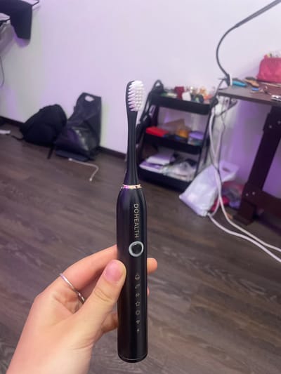 A person's hand, adorned with a thin silver ring, holds a black electric toothbrush. The toothbrush has "DOHEALTH" printed vertically on its body, along with a circular button and several small icons below it. The background suggests an indoor setting with a wooden floor, a desk with items, and a wall illuminated by purple light. The image does not provide any information about the location of Romanovka, Russia.