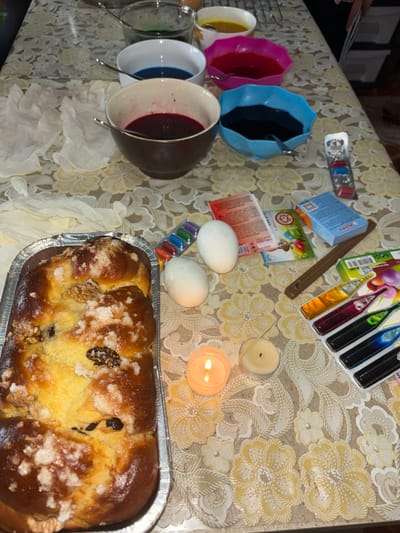 The image shows a tabletop scene, likely indoors, with various items related to Easter preparations. There are several bowls containing liquids in different colors, along with a container of eggs, suggesting the dyeing of Easter eggs. The presence of colored dye tablets and liquid dye further supports this. Two lit candles are visible, providing a warm glow to the scene. A freshly baked sweet bread loaf, likely a traditional Easter bread (Kozunak), is also present. The tablecloth has a floral pattern. Given the presence of Easter-related items and the location in Makariopolsko, Bulgaria, it is probable that this scene depicts preparations for the Easter holiday.