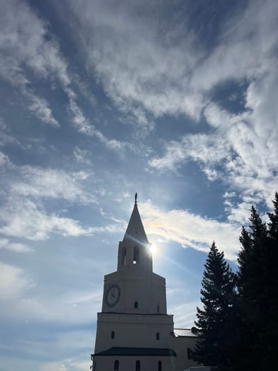 The image displays a low-angle view of a prominent, light-colored architectural tower against a bright blue sky filled with scattered white clouds. The tower, which appears to be a clock tower, features a visible clock face with indistinct Roman numerals. A tapering spire, topped with a finial, extends from the tower's peak. The sun is positioned directly behind the spire and the upper section of the tower, creating a strong sunburst effect and bright lens flare, indicating a sunny daytime scene.
Dark green foliage, likely from evergreen trees, is visible silhouetted against the sky along the top and upper right edges of the frame. The tower's surfaces are well-lit, showcasing its structural details. No people are visible, and no specific activities are taking place. The only visible text is the largely indistinguishable Roman numerals on the clock face. The provided location context for this media is Песчаные Ковали, Russia.