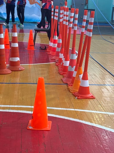 The scene depicts an indoor sports hall in Tayshet, Russia, featuring a wooden floor with distinct red and yellow/orange painted sections, delineated by white and blue lines. In the foreground and midground, a series of approximately twenty orange traffic cones are systematically arranged, some bearing the "TORRES" brand. Interspersed with these are multiple slender orange poles, each marked with white reflective stripes, standing upright from cone bases.
In the background, at least three individuals are partially visible from the waist down. They are dressed in dark trousers and athletic tops, some featuring red, white, and blue patterns, and appear to be wearing harnesses. One individual is positioned on a black folding chair, with the Russian word "СУДЬЯ" (JUDGE) clearly legible on their back. Behind the individuals, a large blue and white banner with graphic designs is displayed against a light blue wall, where blue ropes or cables are also present. The arrangement of cones and poles suggests a structured course or competition.