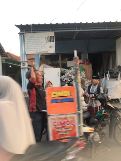 A man in a black and red shirt stands in front of an orange sign. To his right, a young boy wearing a peci hat and a patterned shirt stands near a motorcycle. A woman wearing a headscarf is partially visible in the foreground on the left. The background shows a building with a sign that lists services such as photo printing, wedding photography, and computer services. The overall scene appears to be a street in Sentul, Indonesia.