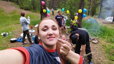 In the city of Orekhovo, Russia, a group of people are gathered in a wooded area for an outdoor event, possibly a camping trip or picnic. In the foreground, a woman is taking a selfie, smiling and pointing towards the camera. She is wearing a dark shirt with red trim. Behind her, several other individuals are visible. One person is standing, appearing to hold something in their hand, while another is sitting on a bench. In the background, tents are set up, suggesting overnight stays. Balloons in various colors are tied to trees, indicating a festive atmosphere. A small fire is being tended to, with smoke rising from it. The environment is characterized by tall trees, green grass, and a natural, somewhat wild setting, likely a forest or park. The overall scene suggests a recreational gathering with a celebratory mood.