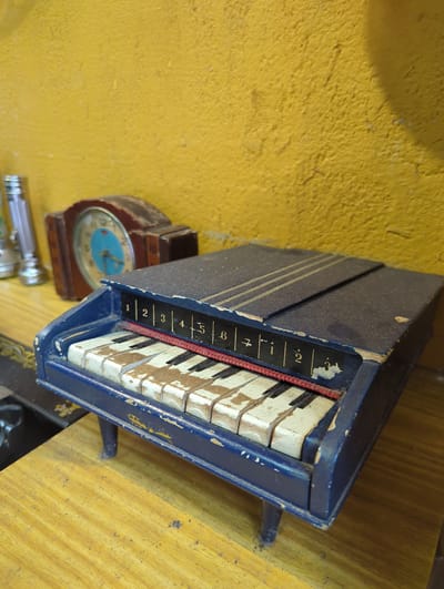 A vintage toy piano with a blue wooden casing and white and black keys sits on a yellow wooden surface. The piano has a worn appearance, with chips in the paint and visible dust. Next to the piano is an antique alarm clock with a brown wooden frame and a blue clock face. The clock's hands are visible, pointing to approximately 3:50. Below the clock and piano is a silver metallic object that appears to be a flashlight. The background is a textured yellow wall. The scene is illuminated by natural light, suggesting it might be daytime. The overall impression is one of nostalgia and the passage of time. There is some faint black text on the side of the piano, partially obscured by wear, with numbers 1 through 6 visible.