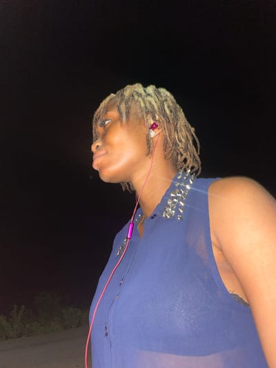 A person with blonde dreadlocks is wearing a blue top with silver spikes. The person has pink headphones in their ear. The background is dark. The image may have been taken in Ogbe, Nigeria.