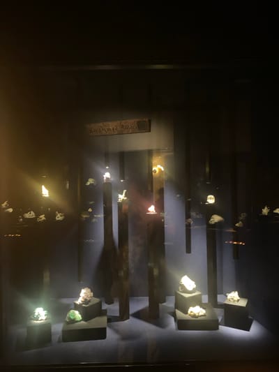 The image shows a display case of minerals, likely in a museum. The display features a series of dark, rectangular shelves holding various mineral specimens, illuminated by spotlights. The specimens appear to be crystals and mineral formations with a range of colors, including white, gold, and green. A plaque is visible with the inscription "Grenoble Alpes Metropole". The overall environment is dark, emphasizing the illuminated minerals.