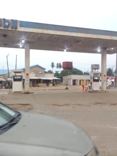 The image shows a gas station with a canopy covering two gas pumps, and a single white car in the foreground. The sign on the canopy reads "Mobil". There is a small red water tank in the background, and a few other buildings are visible. Two people are walking towards the gas pumps. The gas station is located in Adanisievbe, Nigeria, at the coordinates 5.949 latitude and 5.695 longitude. The sky is cloudy, and the environment is dusty. There is a red plastic chair sitting in the middle of the gas station.