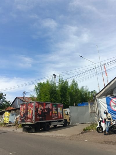 A medium-sized truck with a large advertisement for "So Fresh" is parked on the side of a street. The ad features people and product images. A person is standing near a parked motorbike next to a white building covered in a draped banner. In the background are trees, a utility pole, and a building with a rusty roof. The sky above is blue with scattered clouds.