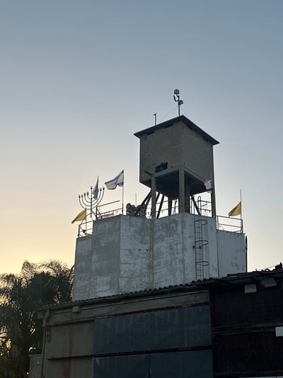 The image displays a multi-tiered tower structure located in Kfar Monash, Israel, captured during either sunrise or sunset, evidenced by the sky's gradient from pale blue to a warm golden light on the left horizon. The base of the tower is a wide, white-painted concrete section exhibiting weathered surfaces and areas of repair. A black metal ladder is affixed vertically to the right side of this lower segment. Above, a narrower, open-sided platform is supported by concrete pillars and angled reinforcement beams. The uppermost section is a smaller, enclosed rectangular structure constructed from light-colored blocks, likely an observation post. An antenna system, featuring multiple dish-like elements on a central pole, is mounted on the roof of this top structure.

Numerous flags are visible: several Israeli flags, characterized by a blue Star of David and blue stripes on a white background, fly from poles attached to both the lower and middle tiers. Additionally, two plain yellow flags are displayed from poles, one on the left and one on the right. A prominent, dark-colored, seven-branched menorah sculpture is positioned on the railing of the lower tier, towards the left. Partial views of lower buildings with corrugated roofs and dark-colored walls are visible in the foreground at the bottom right. Silhouetted palm trees are discernible on the left side against the lighter sky. No individuals are present in the scene.