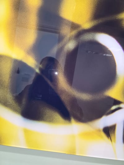 The image captures a close-up, angled view of a reflective surface, likely a framed artwork or screen, situated in Mouscron, Belgium. The surface displays an abstract composition characterized by diffused yellow and dark gray-black areas, overlaid with prominent white, curvilinear, and circular forms. A clear reflection is present on this surface, revealing the upper torso and head of an individual, presumably the photographer, holding a smartphone. The smartphone's screen or flash is visible as a distinct white light source within the reflection. The reflected background indicates an indoor environment with light-colored walls, a dark, bulbous object suspended on the left wall, and a horizontal dark line or shelf on the right wall. The lower edge of the reflective surface is visible, resting on a light grey-green support. Fine scratches are discernible on the reflective material.