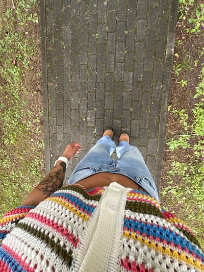 A first-person perspective image shows a person standing on a paved walkway surrounded by grass. The person is wearing blue jeans, brown shoes, and a colorful crocheted cardigan with white buttons. The person's left arm, with a floral tattoo, is visible. The walkway is made of dark grey rectangular pavers arranged in a horizontal pattern. The grass to the sides of the walkway is green with some brown patches. The lighting suggests an overcast day. The location of Duvankul', Russia is not evident in the image.