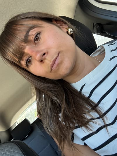 A woman with brown hair and brown eyes is sitting in a car. She is wearing a white shirt with black stripes and a gold earring. The car is parked. The location is in the city of Mougins, France.
