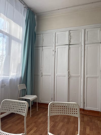 An interior view of a room in Tayshet, Russia, depicts an empty space illuminated by bright daylight from a large window on the left. The room features a light-colored, possibly beige, painted wall and a white ceiling with visible cracks and molding. A reddish-brown wooden laminate floor covers the ground. The window is fitted with a white sheer curtain and a light-teal colored curtain, both hanging from a ceiling-mounted track. Below the window, a white radiator is visible. The entire right wall is occupied by a floor-to-ceiling built-in storage unit, constructed from multiple white-painted wooden panels and doors, some with visible hinges and simple handles. Three white metal chairs with perforated backs and seats are present in the room; one is in the foreground with its back to the viewer, another is positioned closer to the window, and a third is partially visible in the far right foreground. No individuals are present, and no actions or interactions are occurring.