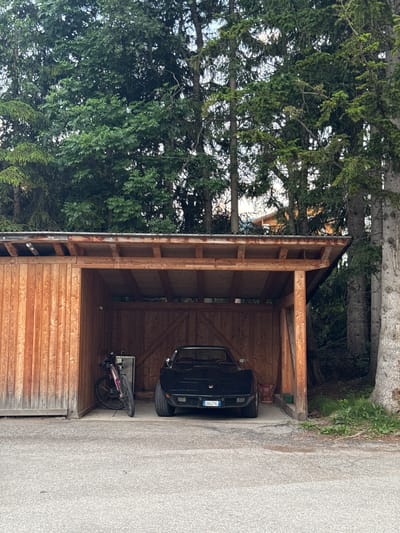 The image depicts a sheltered wooden structure, likely a carport or storage shed, situated outdoors. It is filled with various objects, including a black car, a bicycle, and what appears to be a metal cabinet or drawer unit. The car is parked inside the structure, with its front end visible. The bicycle is leaning against the back wall of the structure, and it has pink accents. The wooden shed has a rustic appearance, with visible grain and natural wood tones.
The background is dominated by lush green trees, suggesting a natural or semi-rural setting. The time of day appears to be daytime, with the lighting indicating it could be late afternoon or early evening, given the angle of the light and shadows. The weather seems fair, with no visible signs of rain or adverse conditions.
A license plate is visible on the black car, displaying the text "ZA647VL". The location context provided, "La Ila - Stern - La Villa, Italy," points to a specific region in Italy known for its mountainous landscape and scenic beauty, which aligns with the presence of trees and the general outdoor feel of the scene. The overall impression is one of a functional storage area integrated into a natural environment.