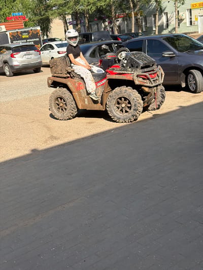 A young woman wearing a white helmet and white pants is sitting on a mud-covered red ATV. The ATV is parked on a dusty surface. In the background, there are cars, buildings, and trees. One of the buildings has a sign that reads "ФРУКТЫ" (Fruits) and another has "КУЛИ" (Kulí). A silver GMC SUV is parked to the left of the ATV, and a dark-colored sedan is parked behind it. The scene appears to be in an outdoor urban environment.
