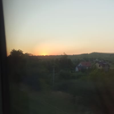 The image captures a sunset over a rural landscape with the sun low on the horizon, casting a warm glow on the sky. In the foreground, a dark, blurry silhouette of trees and vegetation dominates the lower portion of the frame. In the middle ground, a few buildings with red and yellow roofs are visible, partially obscured by the foliage. The background features rolling hills and a distant treeline against the illuminated sky. The overall scene suggests a peaceful, natural environment. The image is taken from a slightly elevated, moving perspective, as indicated by the blur. The location is specified as Zavoloka, Ukraine.