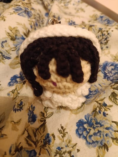 The image shows a crocheted keychain of a doll's head. The doll has skin-toned yarn for its face, black yarn for its hair, and white yarn for a hat or hood. A small purple stitch represents the nose. The doll appears to be wearing a white ruffled collar. A gold keychain loop is attached to the top of the doll's head. The background is a fabric with a floral pattern featuring blue and white flowers with green leaves. The lighting is soft and appears to be indoor lighting. There is no visible text in the image. The image is a close-up shot, focusing entirely on the crocheted doll and the fabric it rests on.