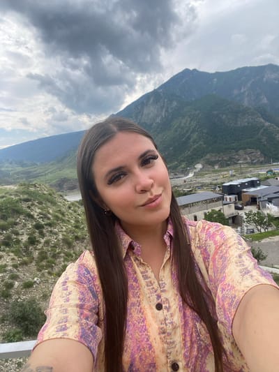 A young woman with long, dark hair is taking a selfie outdoors. She is wearing a light-colored shirt with a multi-colored abstract pattern. Her gaze is directed towards the camera, and she has a slight smile. In the background, a mountainous landscape unfolds under a cloudy sky. The scene appears to be in the vicinity of Chalda, Russia, with a small settlement visible in the mid-ground, nestled amongst the hills. The settlement features modern buildings with dark roofs, and some construction or infrastructure projects are apparent. The natural environment is characterized by rugged terrain with green vegetation covering the slopes.