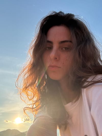A female subject, appearing to be an adult, is captured in a close-up, front-facing photograph. She possesses long, dark, wavy hair, a medium complexion, and a neutral facial expression, gazing directly at the viewer. She is wearing a white garment. The scene is significantly backlit by a low-positioned sun, visible in the lower-left quadrant of the image, casting a bright lens flare that spans the lower-left and central areas of the frame, also illuminating her hair with a radiant, golden-orange hue and producing a subtle prismatic effect within the flare. The background consists of a clear blue sky with minimal, scattered clouds near the horizon, above which the silhouettes of distant mountains or hills are discernible. The depicted environment is in Ban Mae Ping, Thailand.