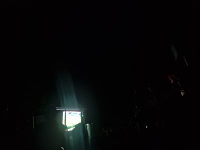 A nighttime outdoor scene in Überlingen, Germany, depicts a gathering of people in front of a large screen or stage. The scene is largely dark, with light rays cutting through the darkness, suggesting a spotlight or projector. A screen on the left side of the image shows bright colorful imagery, possibly a video or performance. Silhouettes of people are visible, some standing and some appearing to be seated. In the upper right of the frame, a person wearing a red head covering is illuminated, possibly looking towards the screen or engaged in an activity. The overall impression is that of an outdoor event or concert.