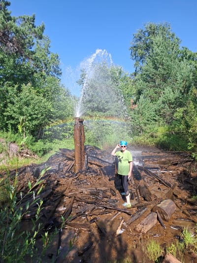 In Razlivnaya, Russia, a person stands in a muddy area with a natural geyser erupting in the background. The geyser is shooting a tall column of water into the air, creating a rainbow. The ground around the geyser is covered in dark, wet debris, possibly tar or mud. The person is wearing a green t-shirt, black pants, blue sunglasses, and a blue bandana. They are holding one hand up to their sunglasses. The scene is surrounded by lush green trees and a clear blue sky. The environment suggests an outdoor, natural setting.