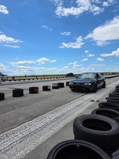 An Audi sedan is parked on a paved track, possibly a drag strip, with stacks of tires lining the sides. The sky is blue with scattered white clouds. In the background, there are fences, trees, and a field. A few people are visible in the distance, near some tents. The scene appears to be outdoors on a sunny day, suggesting a racing or automotive event. The location is specified as Akatsievo, Bulgaria, which is a village in the municipality of Plovdiv, Bulgaria.