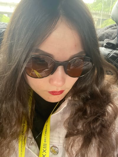 The image is a close-up, angled portrait of a person, likely female, with long, dark brown, wavy hair. The person is wearing dark, roundish sunglasses that obscure their eyes, and their lips are painted with bright red lipstick. A small red blemish is visible on their temple.
Around the person's neck is a bright yellow lanyard, which is a prominent feature. Visible Cyrillic text on the lanyard includes "XXIII" and fragments like "ФЕСТИВАЛЬ ТЕА" (suggesting "Festival of Theatres") and "ГОРОВ МАЛЫХ ГОРОДОВ РО" (likely referring to "of Small Towns of Russia"). This text indicates the person is attending the 23rd Festival of Theatres of Small Towns of Russia.
The person is wearing a light-colored jacket, possibly denim or corduroy, with a silver-toned button visible on the right shoulder area, over a dark-colored top. Reflections in the sunglasses show a blurred environment, possibly a hand holding the camera and parts of a light-colored garment. The background, visible in the upper left, consists of blurred green foliage or trees, suggesting an outdoor view through a window, indicating it is daytime. The photo captures a moment during this cultural event, possibly in Naugol'noe, Russia, or by someone attending from there.